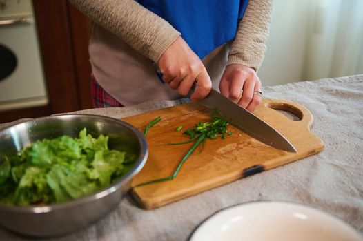 Close-up. Details: Hands of a woman housewife using kitchen knife, chopping young green onion on a wooden cutting board, preparing vegetable raw vegan salad. Healthy eating. Slimming. Dieting concept