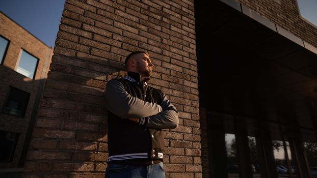 Caucasian bearded man in a bomber jacket leaned against a brick wall