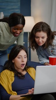 Multi-ethnic friends watching interesting comedy movie on laptop computer relaxing on couch, drinking beer, having fun. Group of mixed race people spending time together during night party
