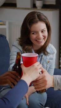 Multi cultural friends cheering beer bottles during wekeend party laughing while relaxing on sofa late at night. Group of mixed race people hanging out enjoying time spending together