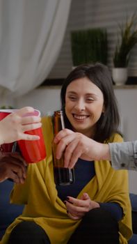 Mixed race friends chatting during night party while sitting on sofa in living room drinking beer having fun. Group of multi-ethnic people hanging out enjoying time spending together