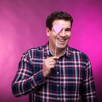 Funny adult man covering his right eye with a pink heart. Studio photo