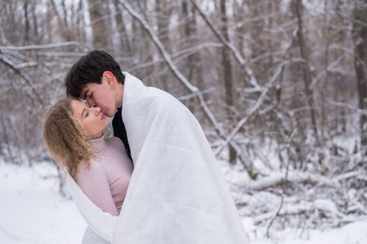 A young couple walks in the park in winter. The guy and the girl are kissing wrapped in a white blanket outdoors