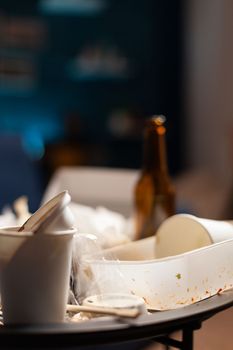 Empty dirty food cans and beer bottles left on unorganized table covered in leftovers garbage and messy trash. Stinky unhealthy rubbish in untidy chaotic living room space. Close up.