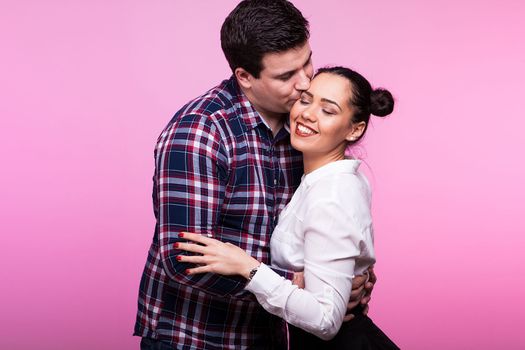 Happy husband kissing his wife on the cheek in studio on pink background