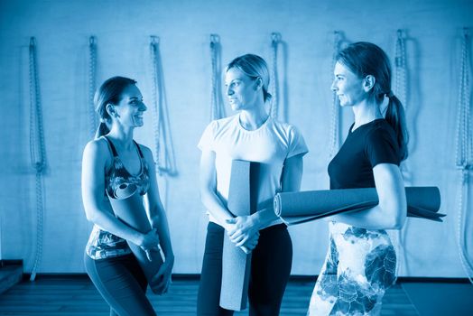 Group of female friends in sportswear smiling together while standing in a gym after yoga workout. Women standing by a wall with exercise mat.