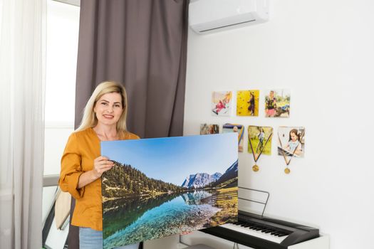 Canvas print with gallery wrap interior decor. Woman hangs landscape photography on white wall. Hands holding photo canvas print.
