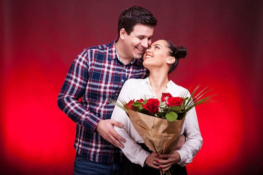 Husband embracing her wife while she holds a roses bouquet in hands on red background in studio photo