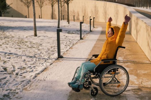 Happy woman in wheelchair happily pulls her hands up while walking in winter park