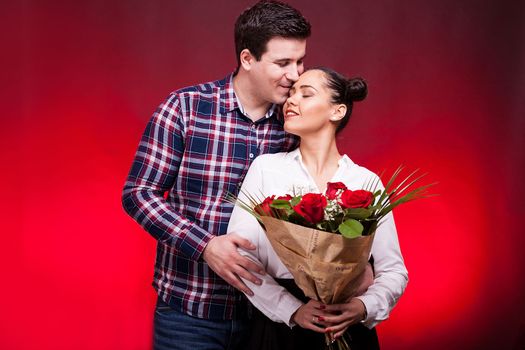 Man embracing her wife while she holds a roses bouquet in hands on red background in studio photo