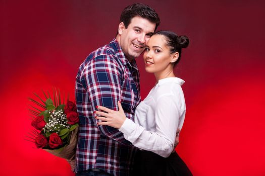 Man with a roses bouquet at his back on a first date embracing a gorgeous girl. Red background and studio photo