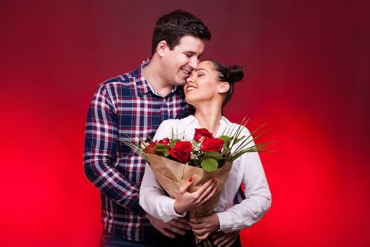 Man embracing her wife and kissing her on the forehead while she holds a roses bouquet in hands on red background in studio photo