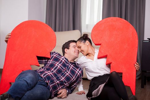 Happy couple holding two parts of one big heart lying on the floor