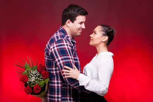 Man with a roses bouquet at his back on a first date with a gorgeous girl looking at her and smiling. Red background and studio photo