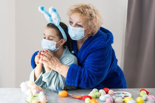 grandmother and granddaughter in medical masks are painting Easter eggs for the holiday. Happy easter family quarantined coronavirus. Happy at home