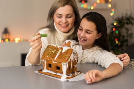 mother and daughter decorating gingerbread house. Beautiful living room with lights. Happy family celebrating holiday together