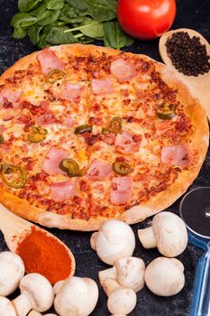 Close up of pizza surrounded with ingredients which it is made from on dark wooden table