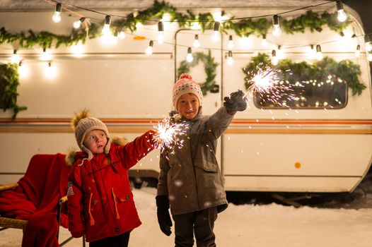 Caucasian red-haired boys hold sparklers by the trailer. Two brothers are celebrating Christmas on a trip