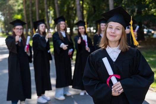 Group of happy students in graduation gowns outdoors. A young girl with a diploma in her hands in the foreground