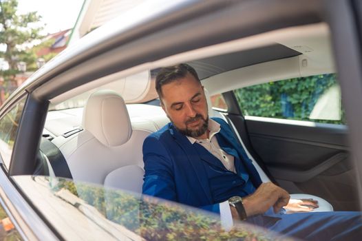 A caucasian man in a blue suit looks at his watch while sitting in the back seat of a car. Business class passenger