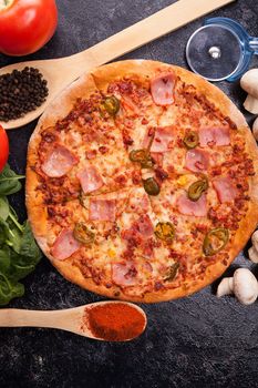 Top view on hot pizza next to ingredients which it is made of on dark wooden board