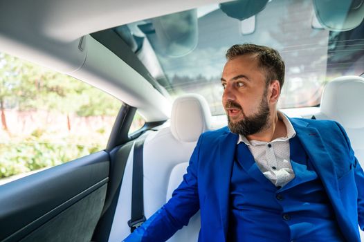 A caucasian man in a blue suit looks out the open window while sitting in the back seat of a car. Business class passenger