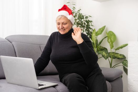 Smiling senior woman wearing Santa hat at Christmas time typing on laptop from home getting in touch with family or friends