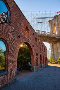 Image of Brick building with arches and golden sun peeking through next to stunning iconic Brooklyn Bridge in New York City