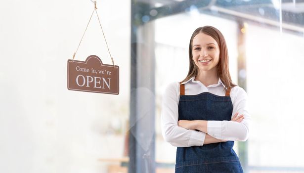 Portrait of a happy waitress standing at restaurant entrance. Portrait of mature business womanattend new customers in her coffee shop. Happy woman owner showing open sign in her small business shop...