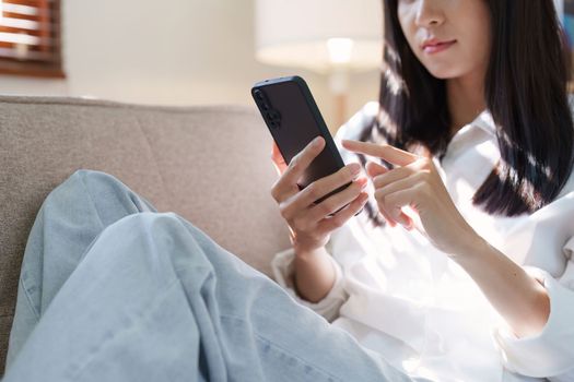 Smiling pretty asian young woman wearing casual clothes using cell phone sitting on sofa at home.