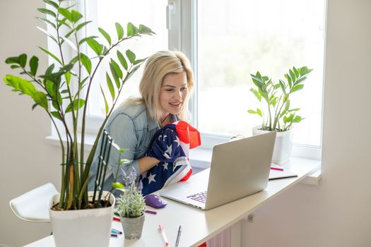 Happy woman employee sitting wrapped in USA flag, shouting for joy in office workplace, celebrating labor day or US Independence day. Indoor studio studio shot isolated on yellow background.