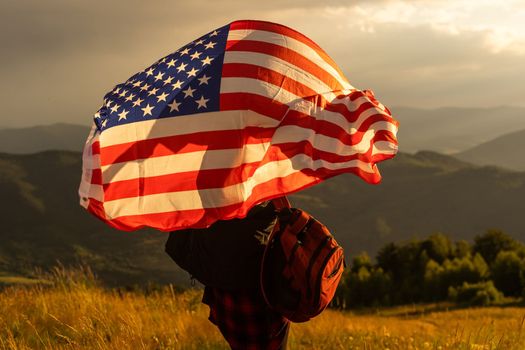 Young happy american woman with long hair holding waving on wind USA national flag on her sholders relaxing outdoors enjoying warm summer day.