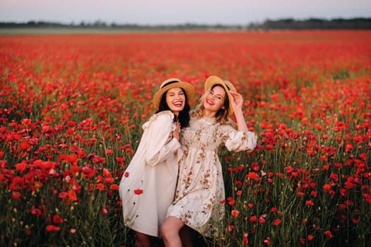 Two girlfriends in dresses and a hat in a poppy field in summer at sunset.