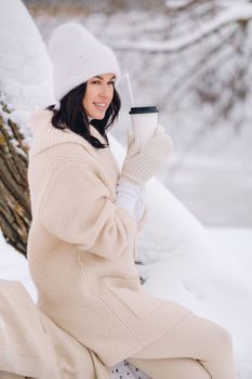 A beautiful girl with a beige cardigan and a white hat enjoying drinking tea in a snowy winter forest near a lake.