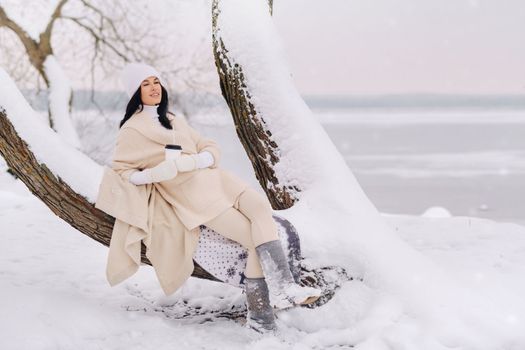 A beautiful girl with a beige cardigan and a white hat enjoying drinking tea in a snowy winter forest near a lake.