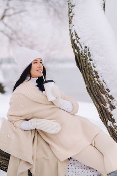 A beautiful girl with a beige cardigan and a white hat enjoying drinking tea in a snowy winter forest near a lake.