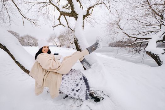 A beautiful girl with a beige cardigan and a white hat enjoying drinking tea in a snowy winter forest near a lake.