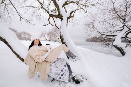 A beautiful girl with a beige cardigan and a white hat enjoying drinking tea in a snowy winter forest near a lake.