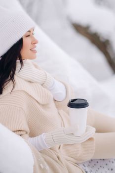 A beautiful girl with a beige cardigan and a white hat enjoying drinking tea in a snowy winter forest near a lake.