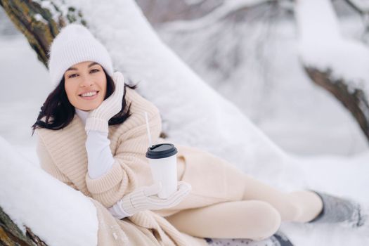 A beautiful girl with a beige cardigan and a white hat enjoying drinking tea in a snowy winter forest near a lake.