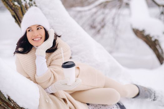 A beautiful girl with a beige cardigan and a white hat enjoying drinking tea in a snowy winter forest near a lake.