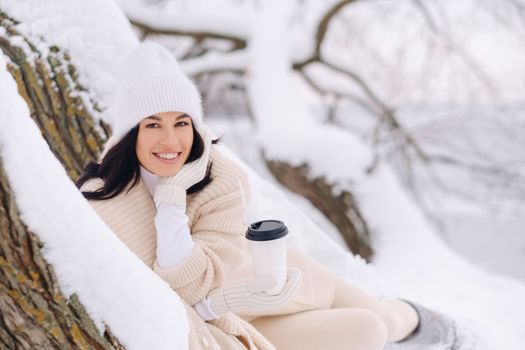 A beautiful girl with a beige cardigan and a white hat enjoying drinking tea in a snowy winter forest near a lake.