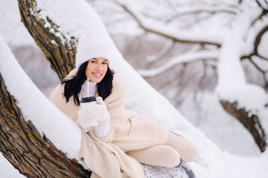 A beautiful girl with a beige cardigan and a white hat enjoying drinking tea in a snowy winter forest near a lake.