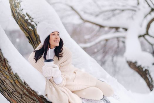 A beautiful girl with a beige cardigan and a white hat enjoying drinking tea in a snowy winter forest near a lake.