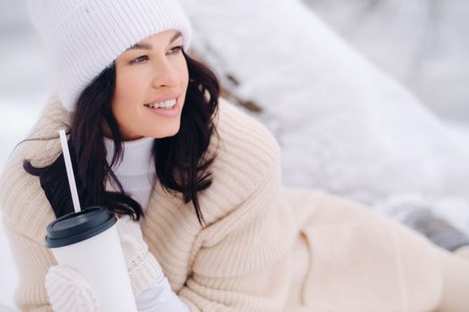 A beautiful girl with a beige cardigan and a white hat enjoying drinking tea in a snowy winter forest near a lake.