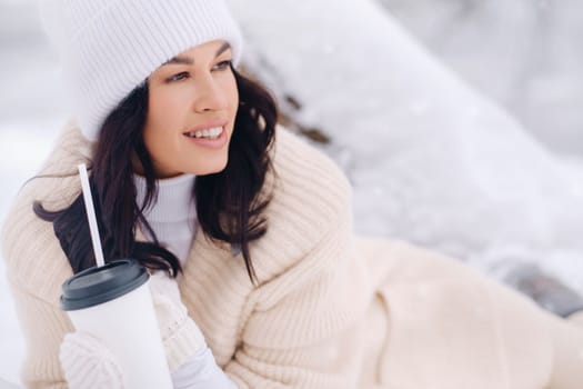 A beautiful girl with a beige cardigan and a white hat enjoying drinking tea in a snowy winter forest near a lake.