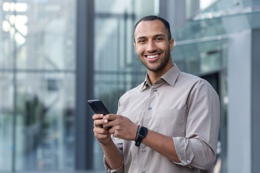 Portrait of African American businessman, man holding phone smiling and looking at camera, office worker in shirt outside modern office building on lunch break.