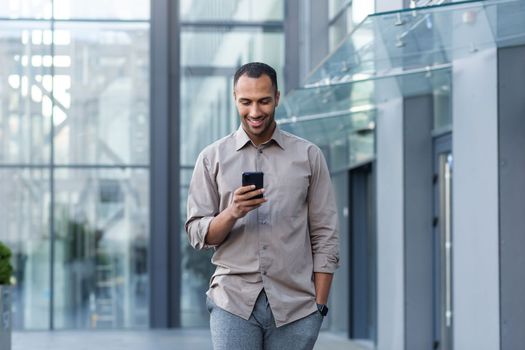 Latin american businessman in casual shirt walking near office building from outside, man smiling and using phone, freelancer typing message and browsing internet pages.
