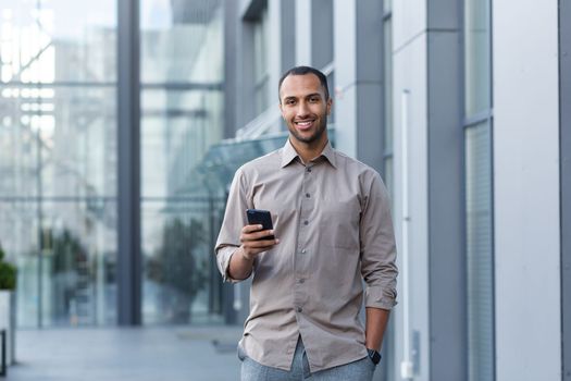 Portrait of African American businessman, man holding phone smiling and looking at camera, office worker in shirt outside modern office building on lunch break.