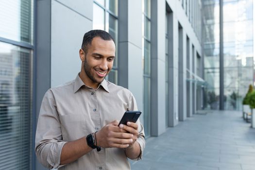 hispanic man outside modern office building using smartphone, businessman in shirt typing message and browsing online pages.